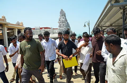 S. Ramesh in a black T-shirt walking across the Tiruchendur temple courtyard with the gopuram behind him, carrying a yellow folder, surrounded by aides