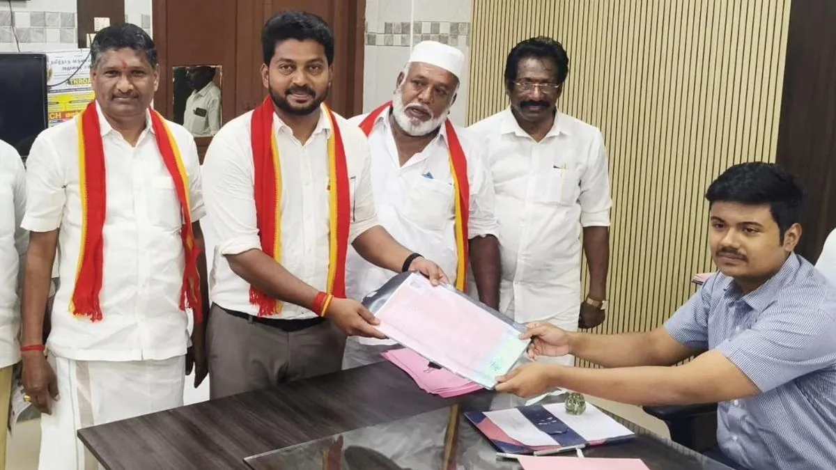 Dr. T. K. Prabhu filing his nomination papers as the TVK candidate for Karaikudi, wearing a white shirt and the red-and-yellow TVK shawl, flanked by senior party figures and a Returning Officer