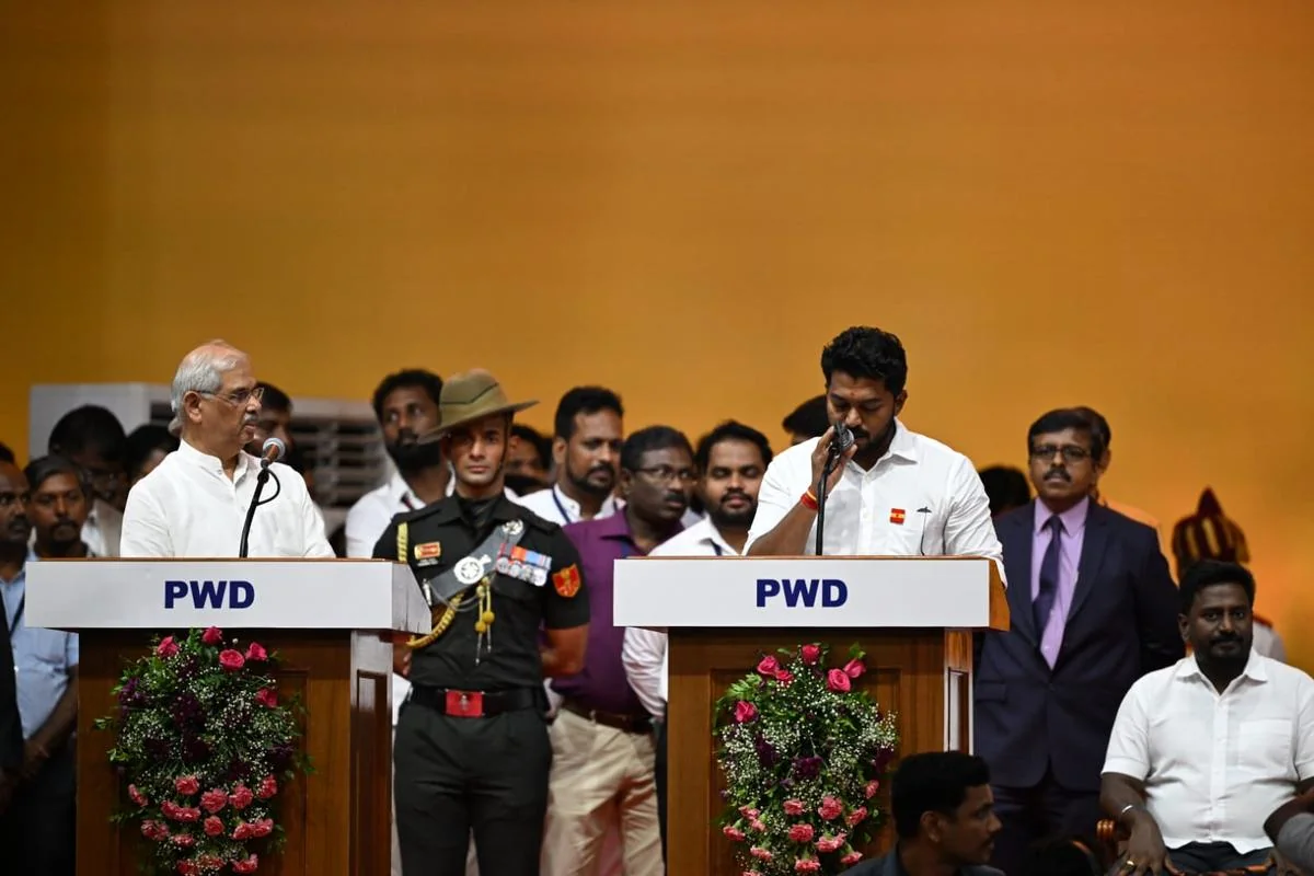 Dr. T. K. Prabhu reading the oath at a podium marked PWD at the Jawaharlal Nehru Indoor Stadium, opposite Governor Rajendra Arlekar, with an Army aide-de-camp standing between the two podiums