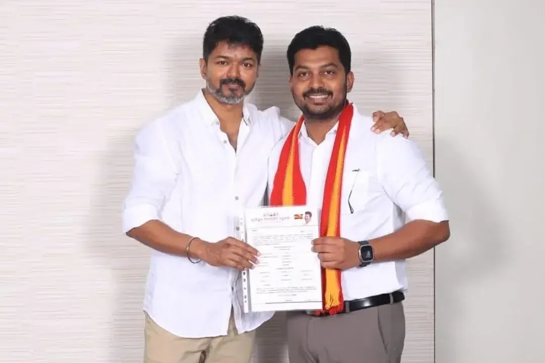 Joseph Vijay and Dr. T. K. Prabhu pose together holding Prabhu's TVK membership paperwork, both in white shirts, Prabhu wearing the red-and-yellow TVK shawl